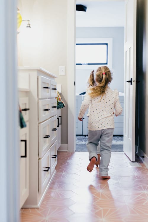 Small children walk through a jack and jill bathroom with redish floors and white cabinets.