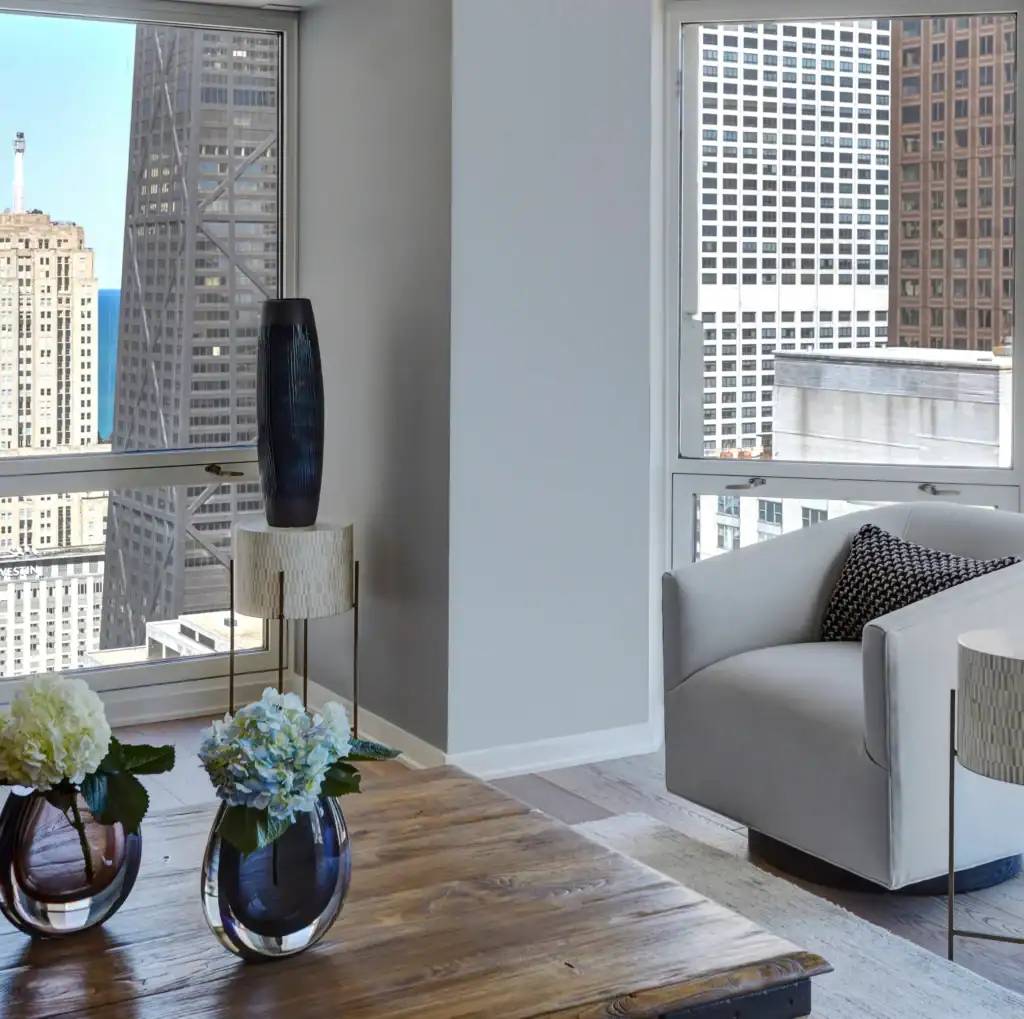 Picture of a bright living room corner with a wooden coffee table, armchair, and tall city buildings outside.